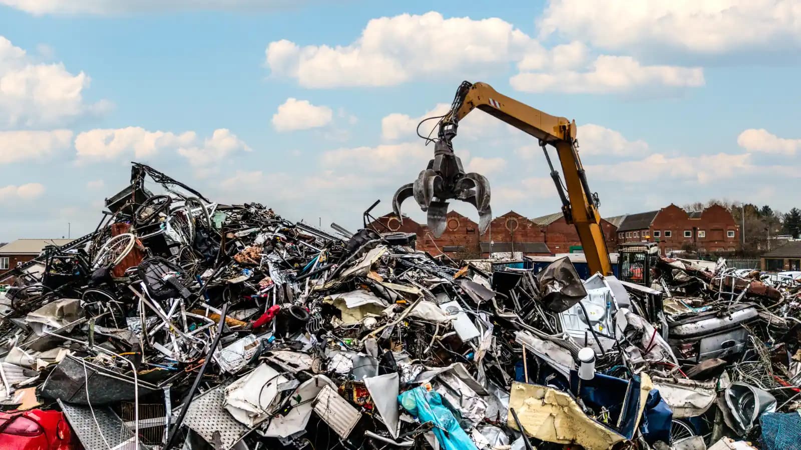 A grapple excavator lifting crushed car parts over a large scrap metal pile at a recycling yard