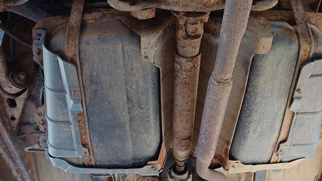 The underside of an older car showing the fuel tank, driveshaft and a rusted exhaust pipe running back