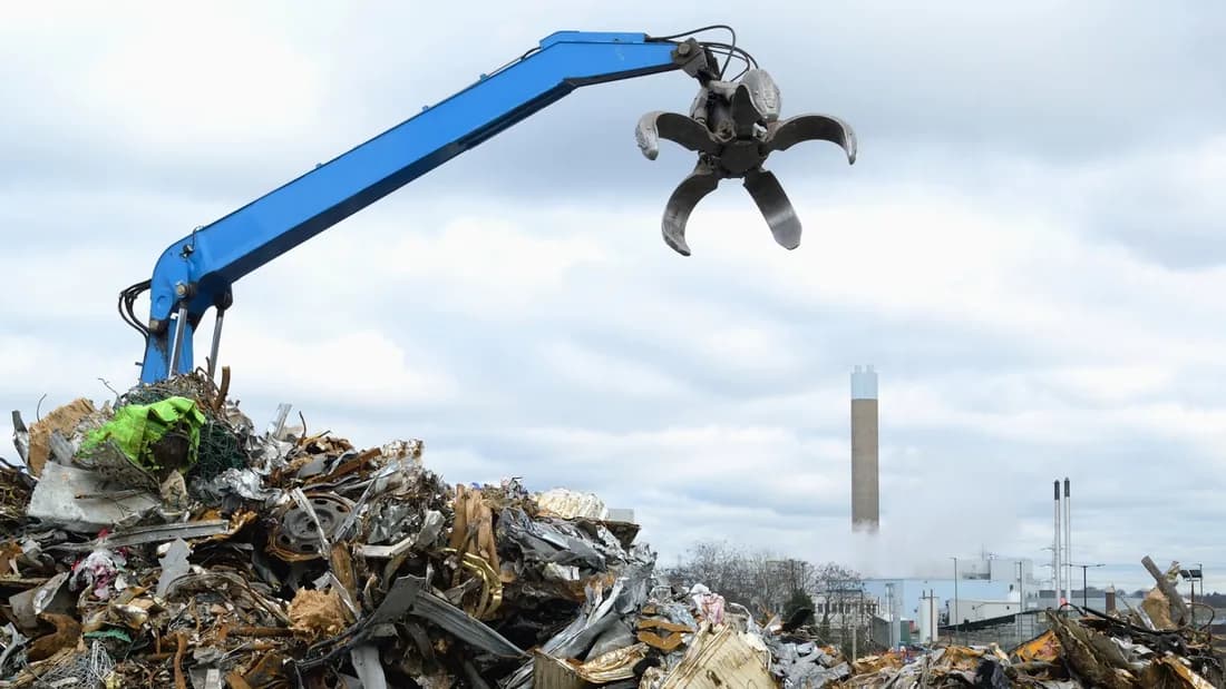 A blue hydraulic claw crane hanging open above a large pile of mixed scrap metal