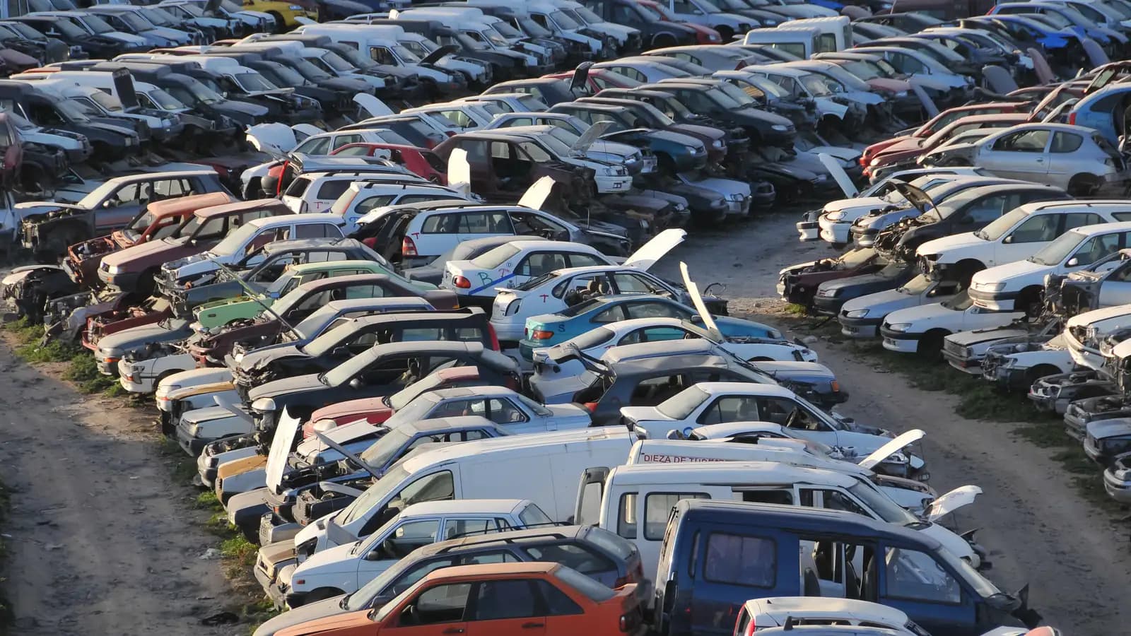 Rows of junk cars and vans parked tightly in a junkyard, many with hoods open and parts removed