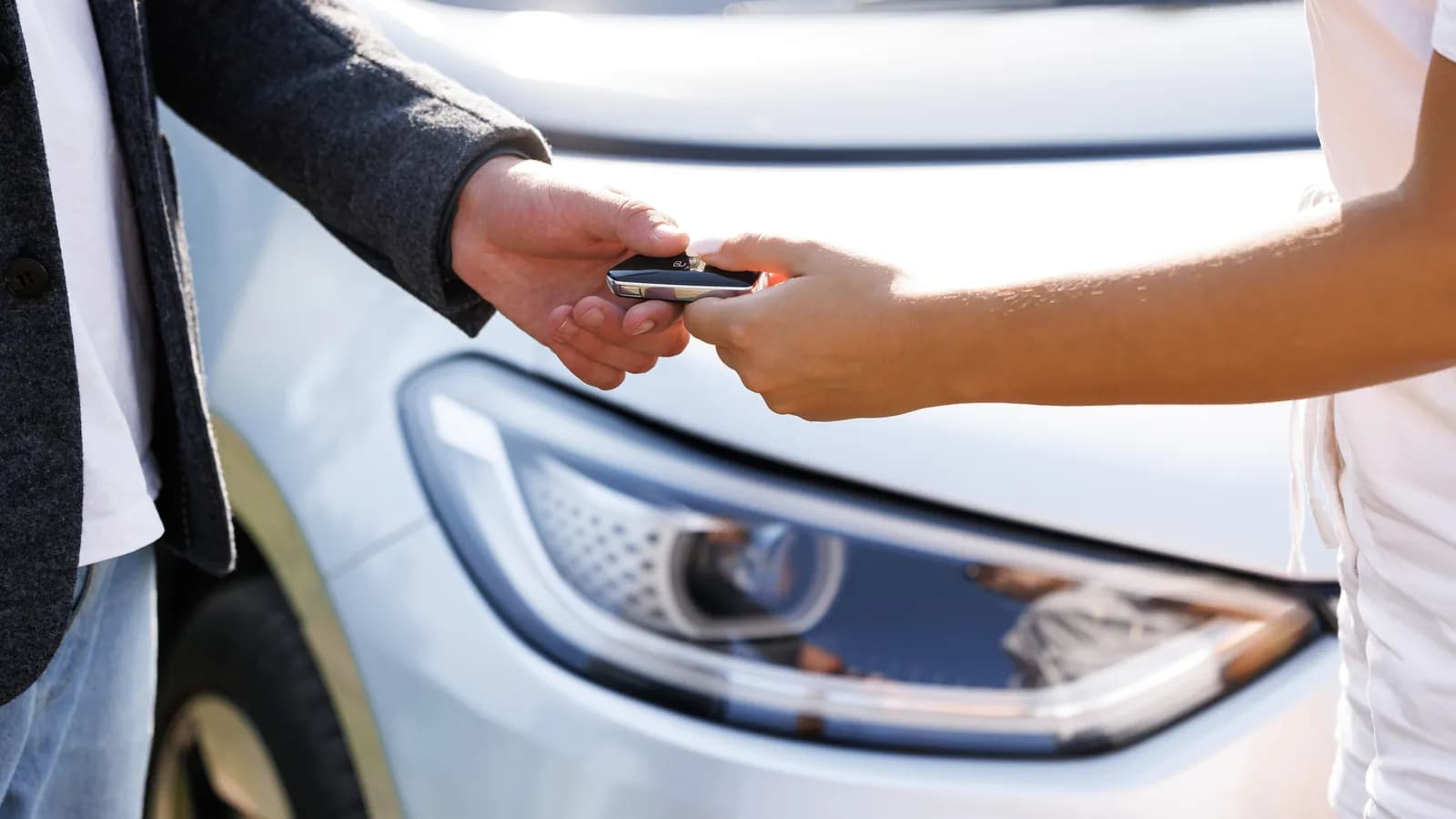 A close view of one person handing a car key to another beside the front of a white car