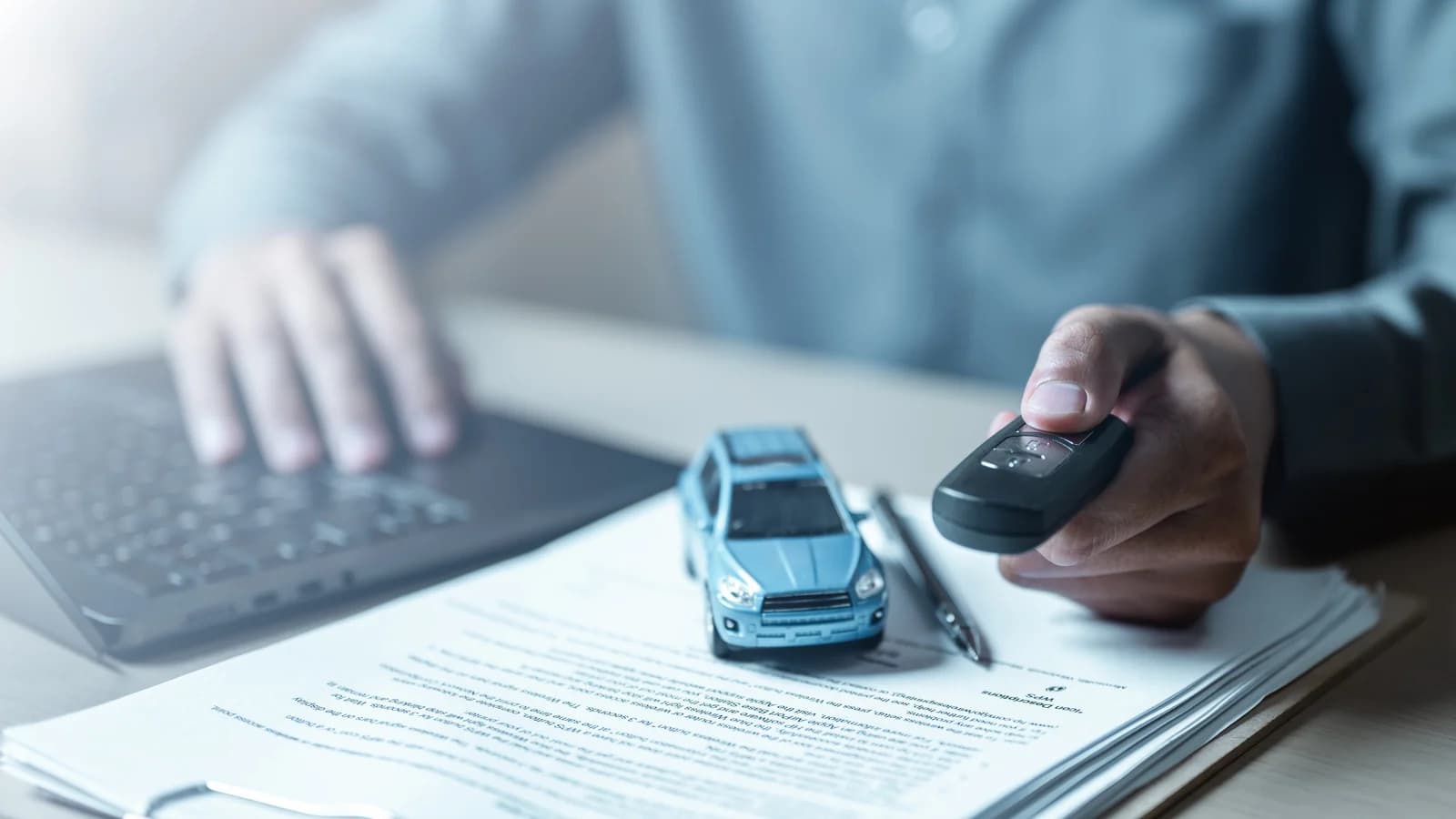 A person typing on a laptop beside a key fob, a toy car and documents: report a sold car to the Nevada DMV with a vehicle resale notification