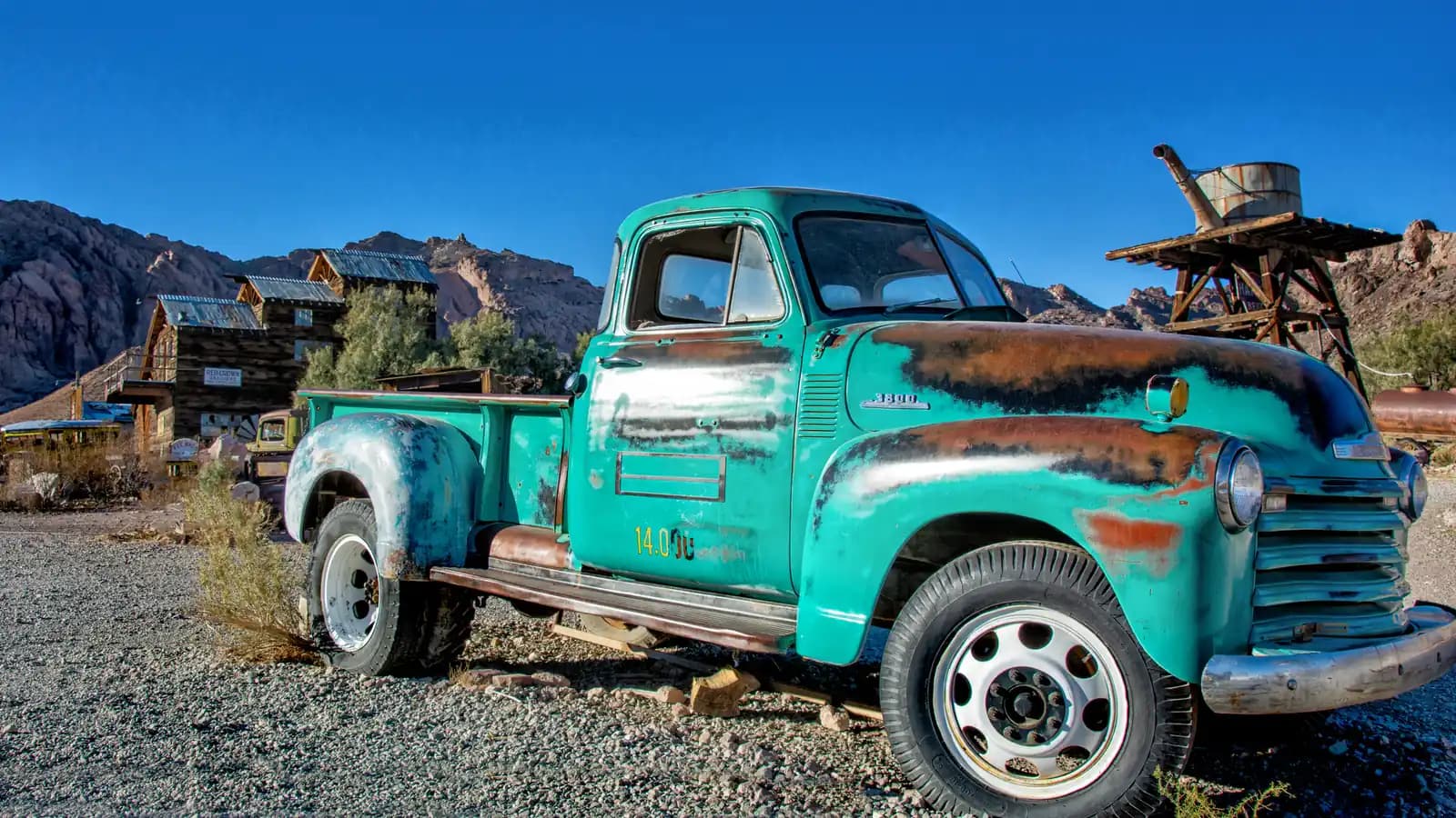 A weathered turquoise vintage pickup truck parked on gravel in front of old wooden buildings and mountains