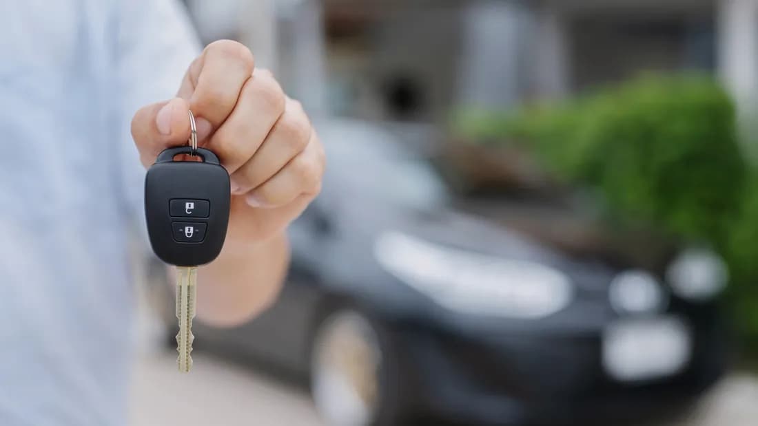 A hand holding a car key in front of a blurred dark car: hand over the key when you sell your car to a buyer in Nevada